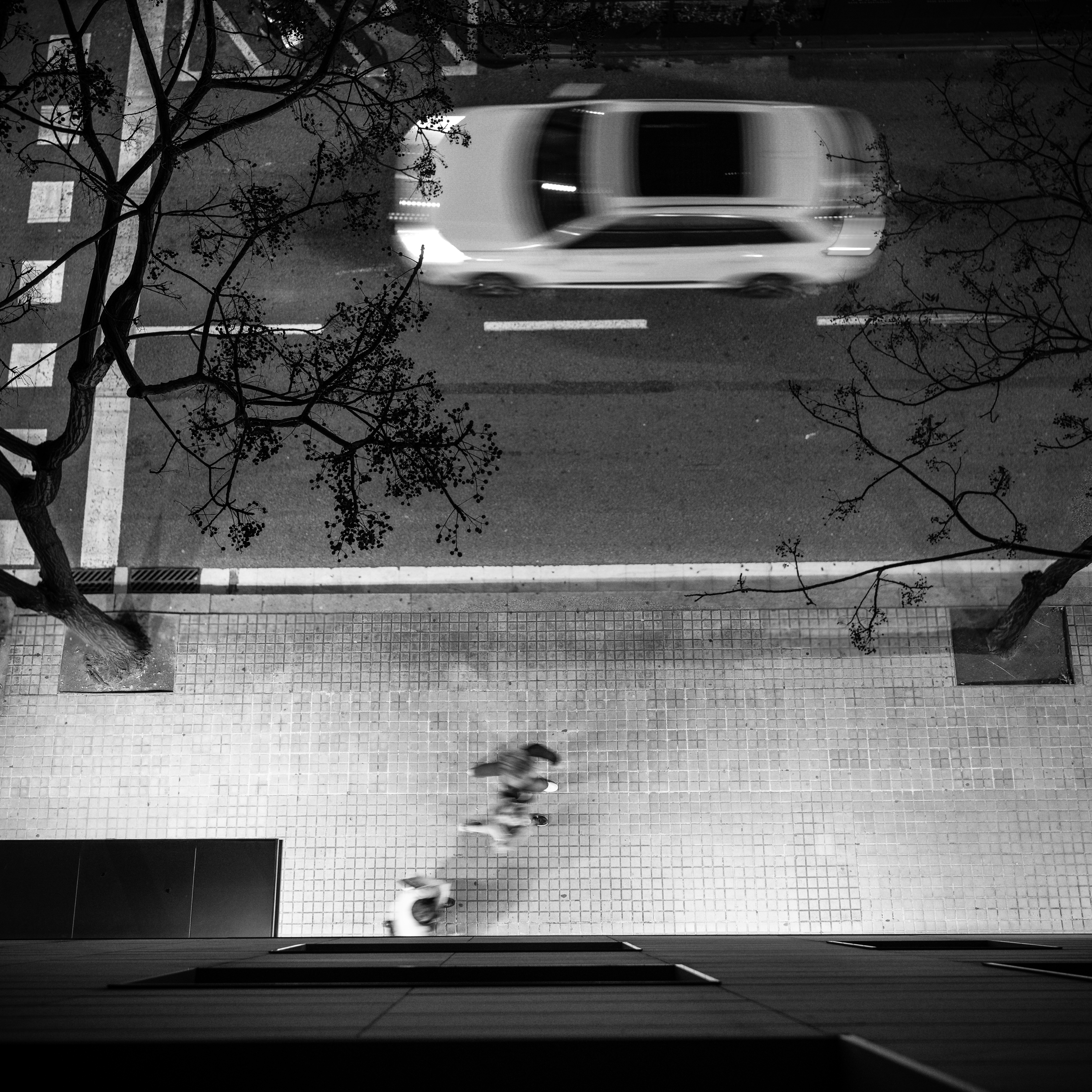 Overhead aerial view of a Carrer de Calvet in Sant Gervasi from my hotel at night, a blurred car and cyclist trailing light streaks below, square paving stones geometric from above