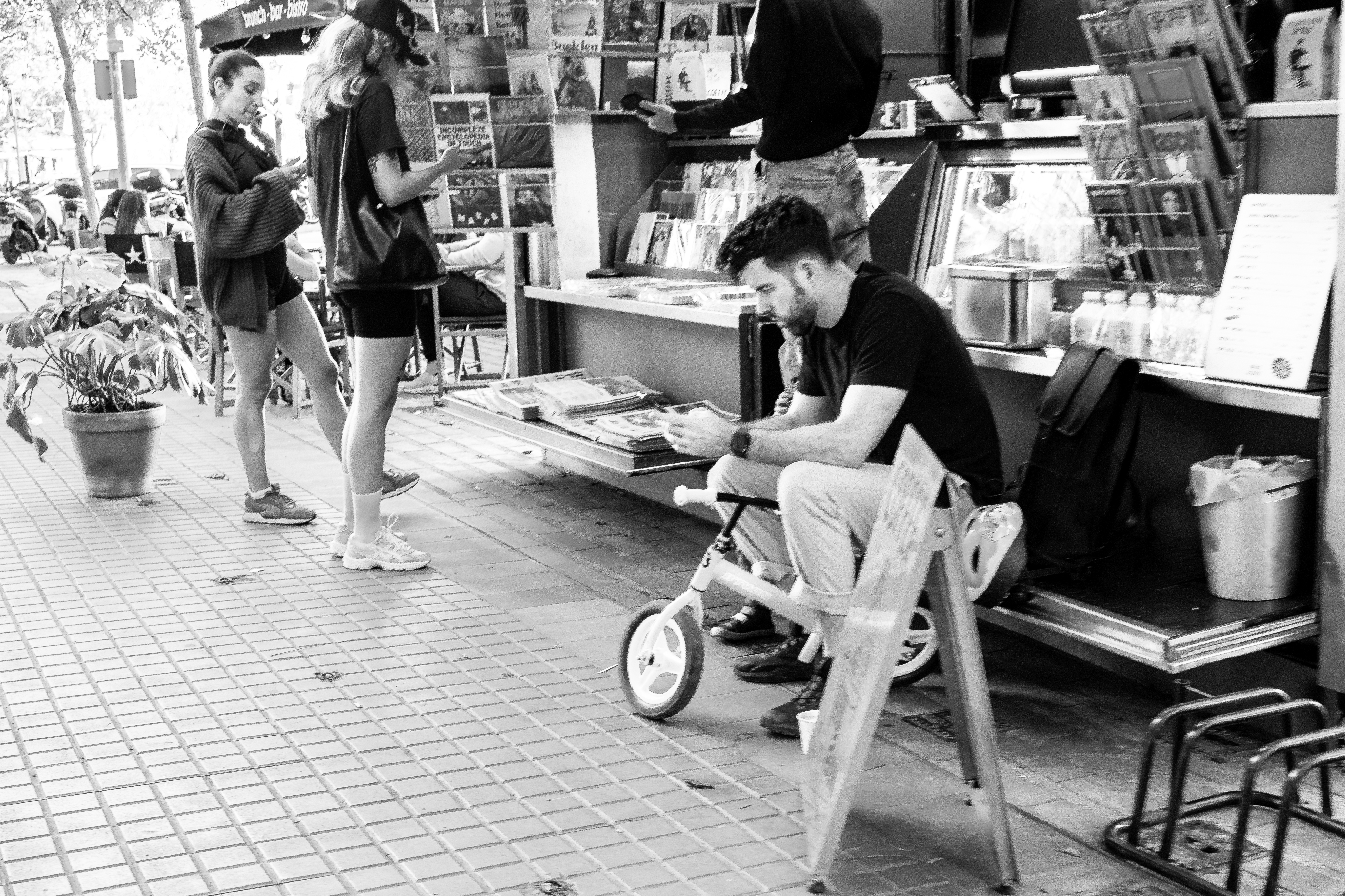 La Rambla book stalls at quiet morning hour, a vendor seated in a folding chair reading beside a child's bike leaning against the stall