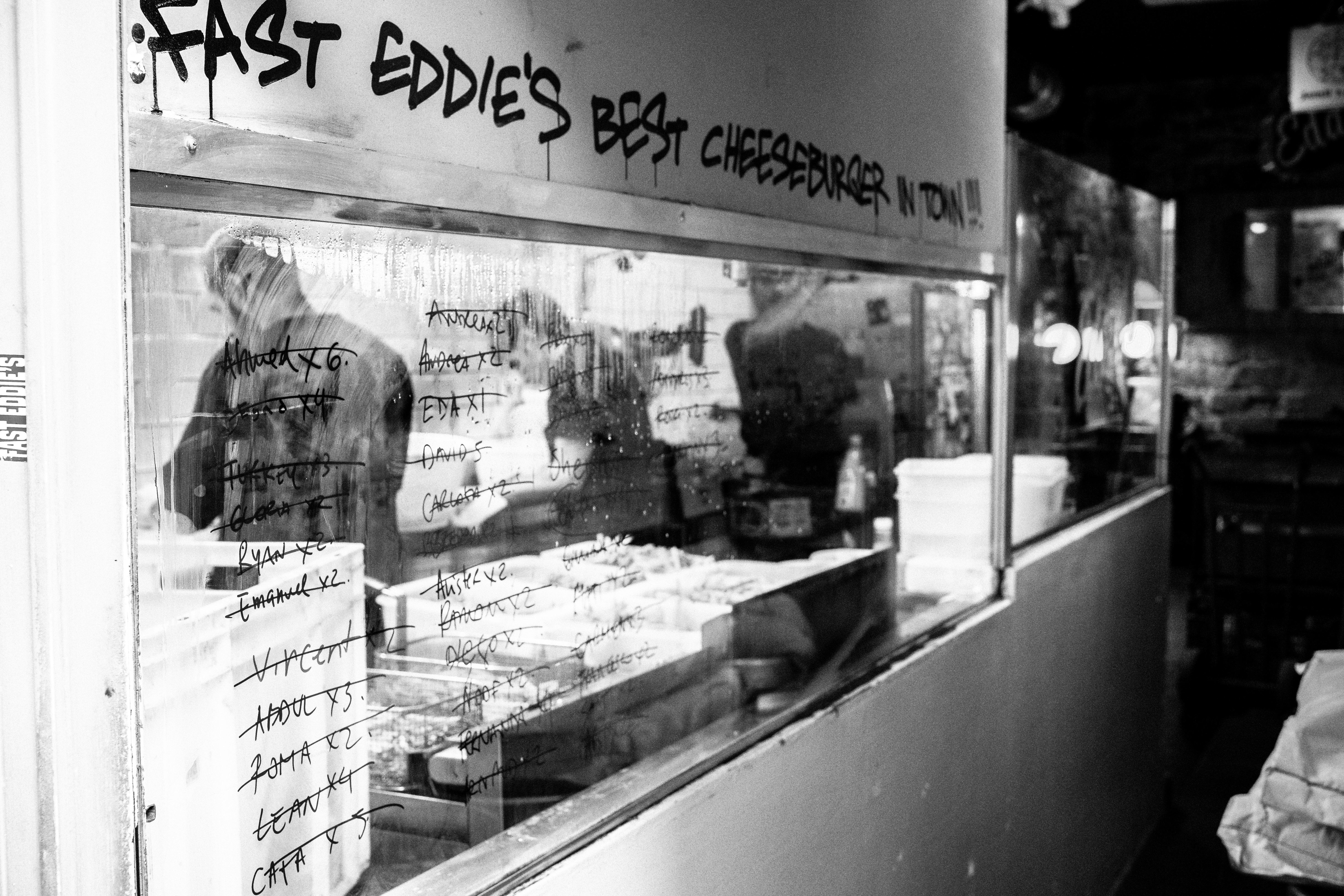 Street-level window reflection in the Gothic Quarter showing the sign for Fast Eddie's Best Cheeseburger overlaid on the stone street and passing pedestrians