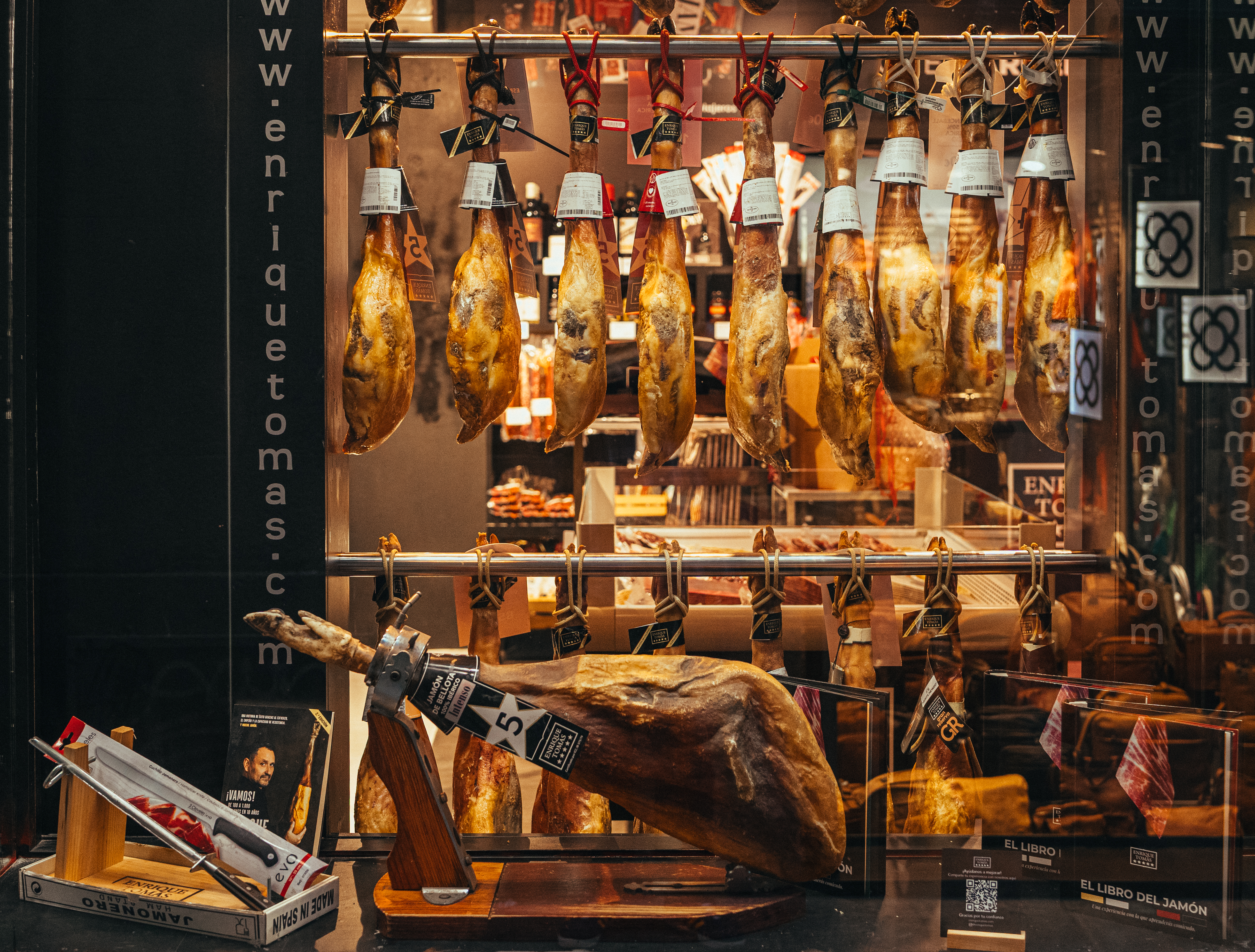 Whole jamón legs hanging in the illuminated window of Enrique Tomás on La Rambla, Barcelona, with a leg mounted on a carving stand in the foreground