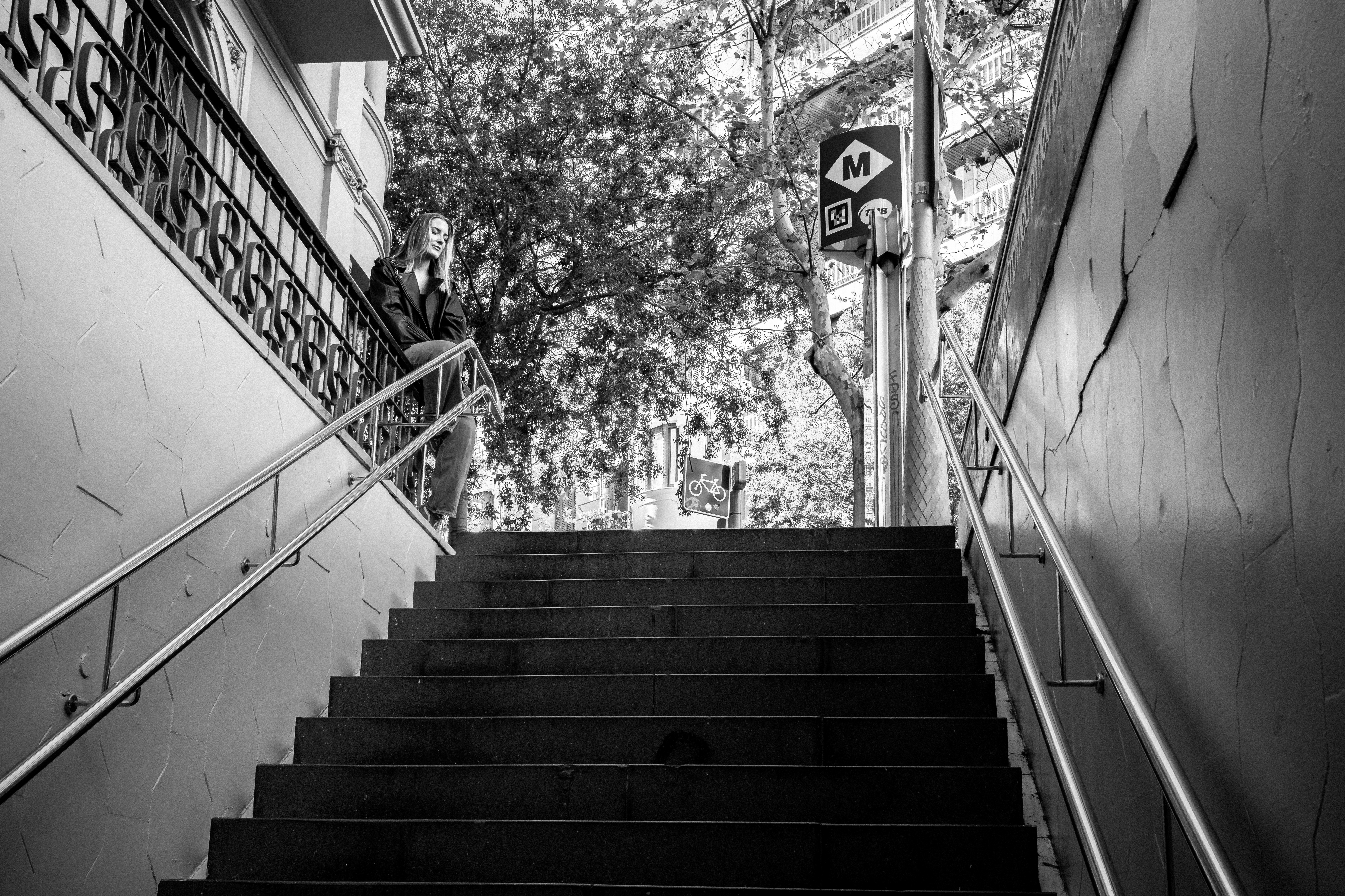 Looking up from below at a Metro staircase, a woman pauses at the top beside the red M sign, light streaming down from street level