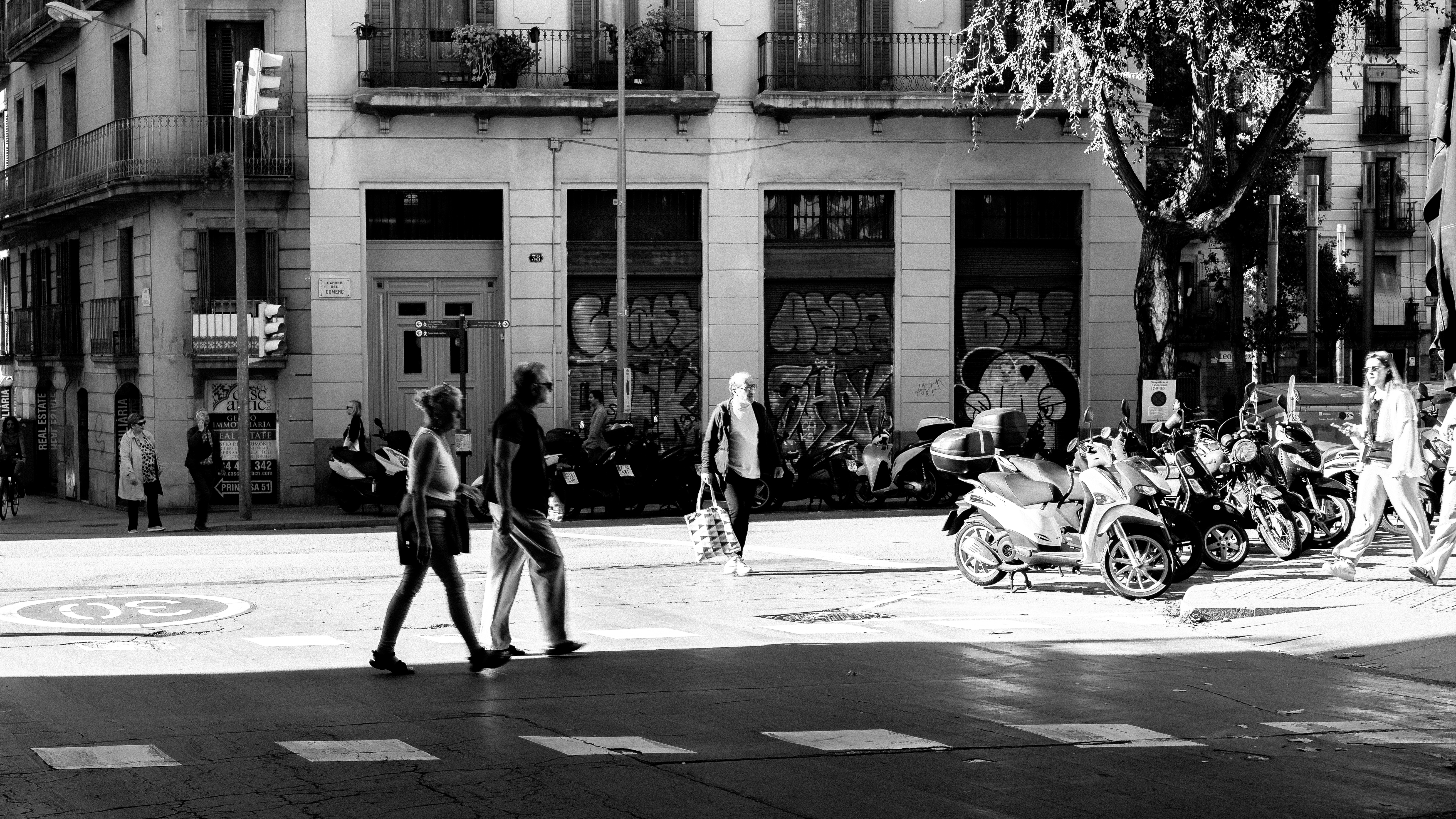 Pedestrians crossing a wide commercial street, silhouetted against bright shopfronts, motorbikes parked on the curb