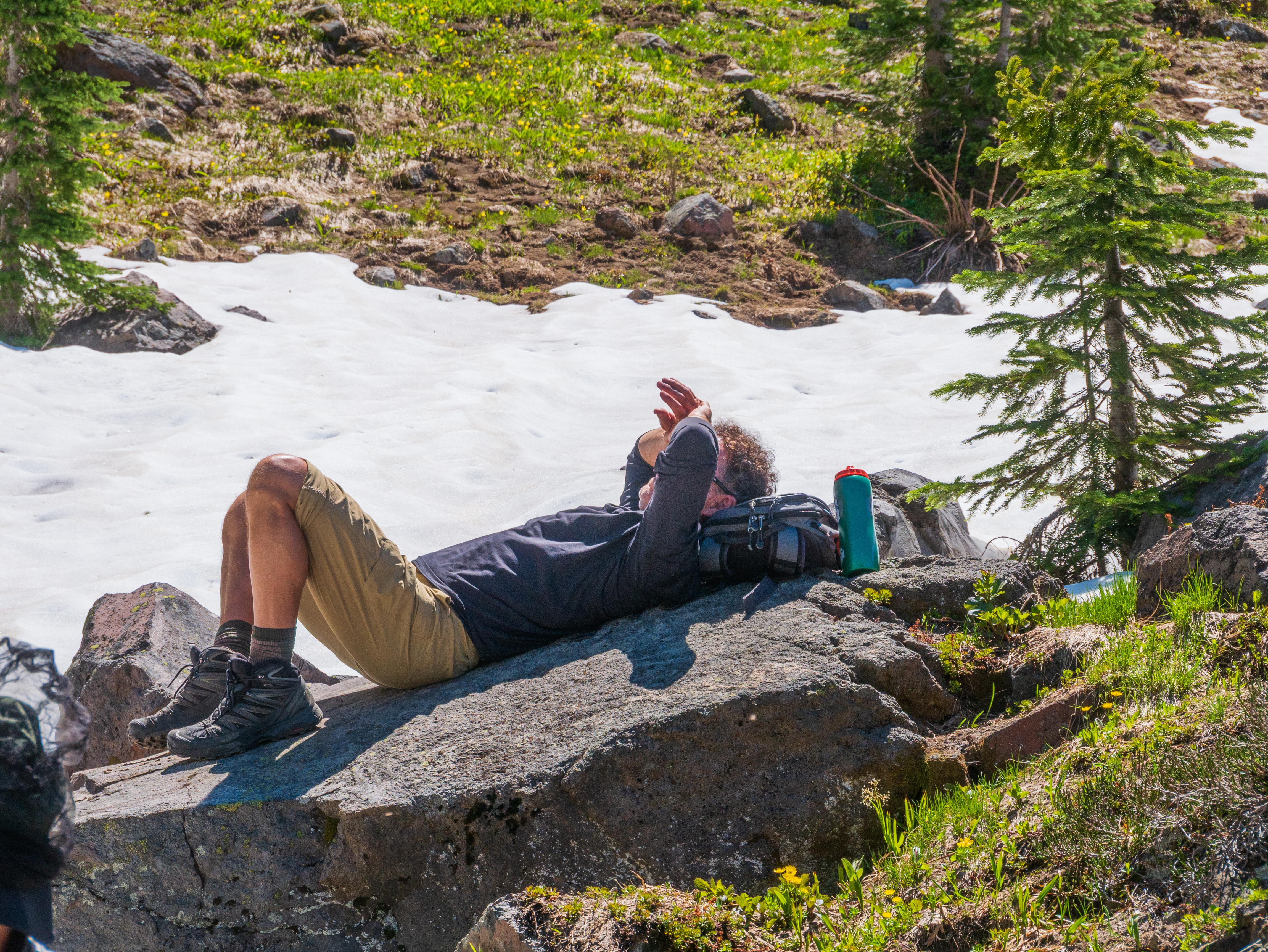 Hiker reclining on a sun-warmed rock in a subalpine meadow beside a broad snowfield, taking a well-earned break in the Goat Rocks Wilderness
