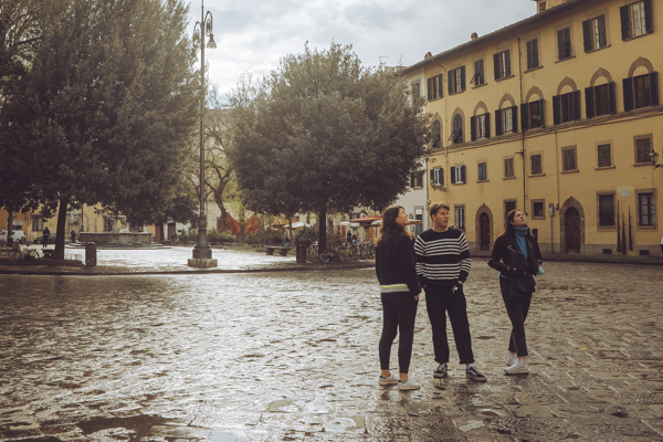Three travelers exploring a wet cobblestone piazza in Florence, ochre buildings and a lamp post behind them