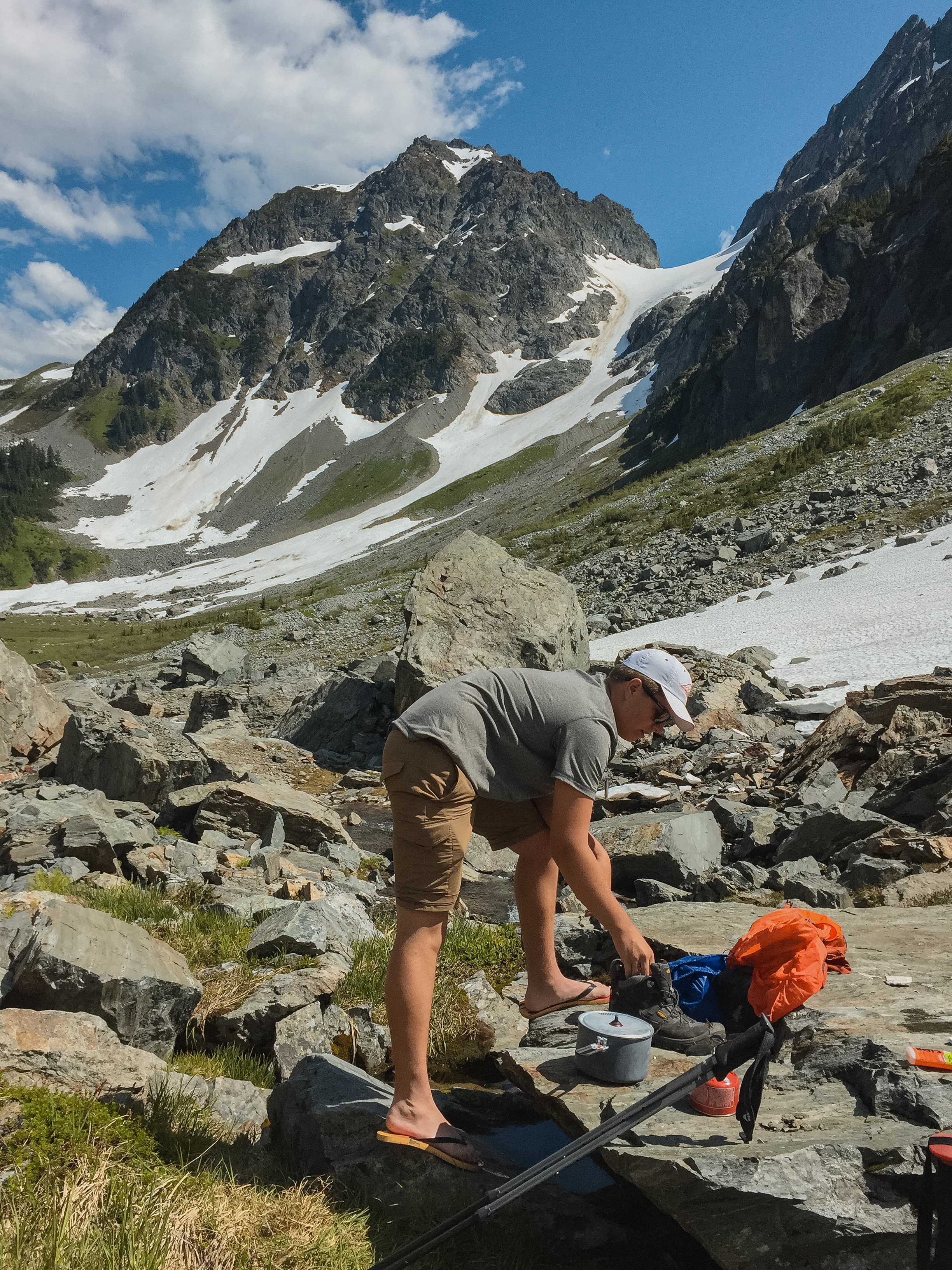 Hiker in flip-flops boiling water at camp in Pelton Basin with a dramatic peak and snowfield rising directly behind