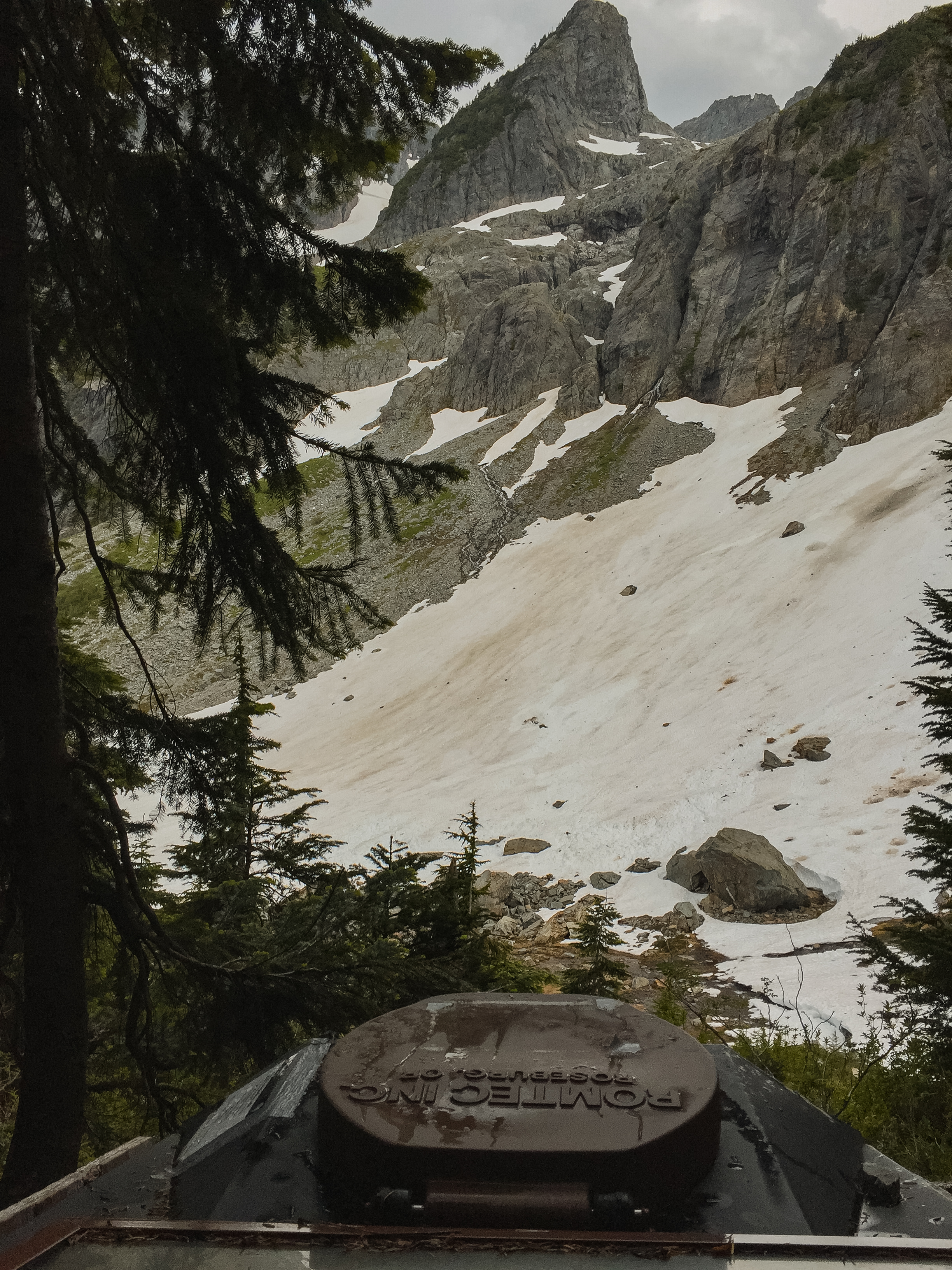 A backcountry camp toilet perched on a ridge with snowfields and dramatic Cascade peaks filling the view behind it