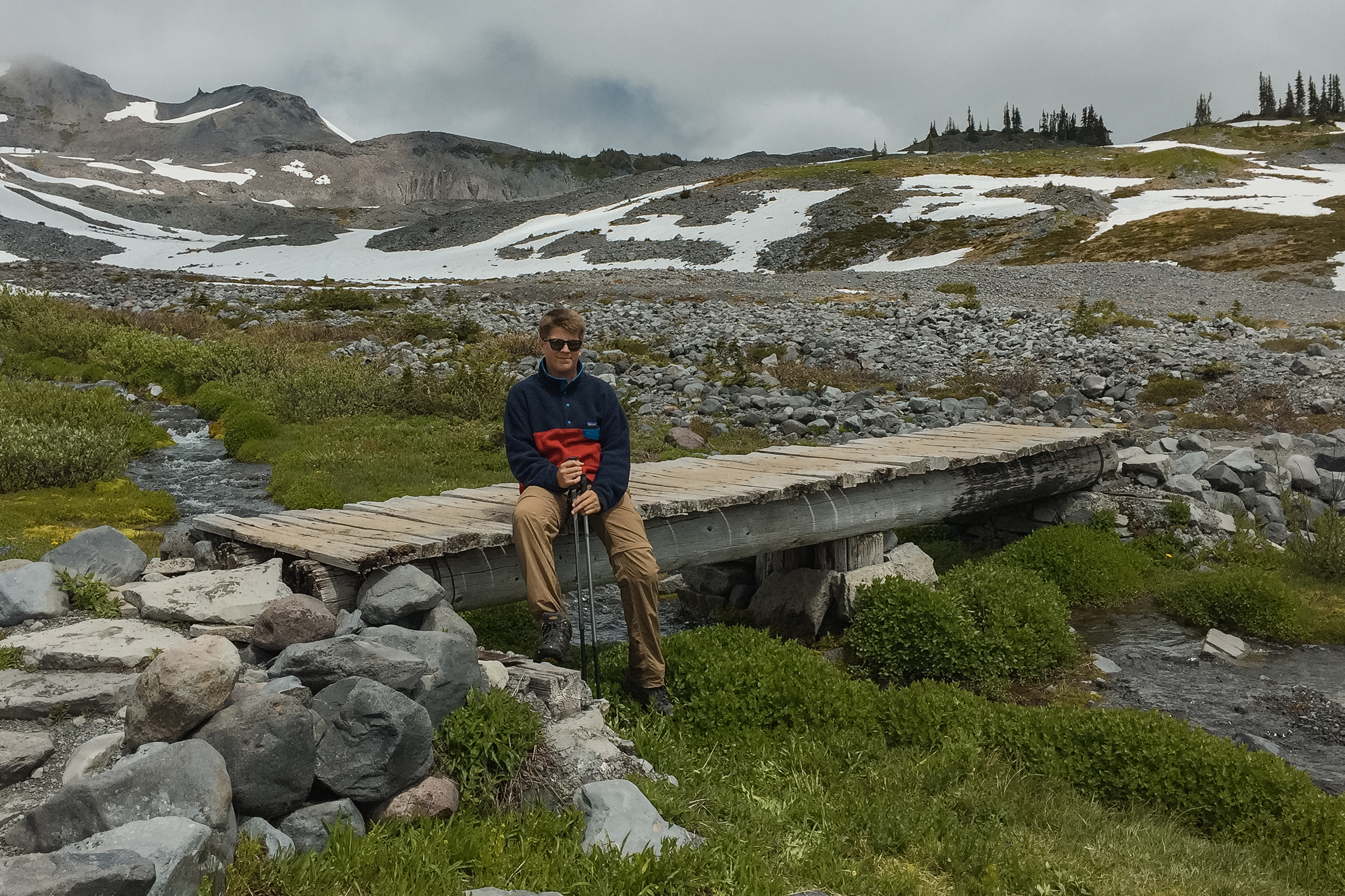 Young hiker sitting on a wooden footbridge in an alpine meadow at Rainier with snowfields and low clouds behind
