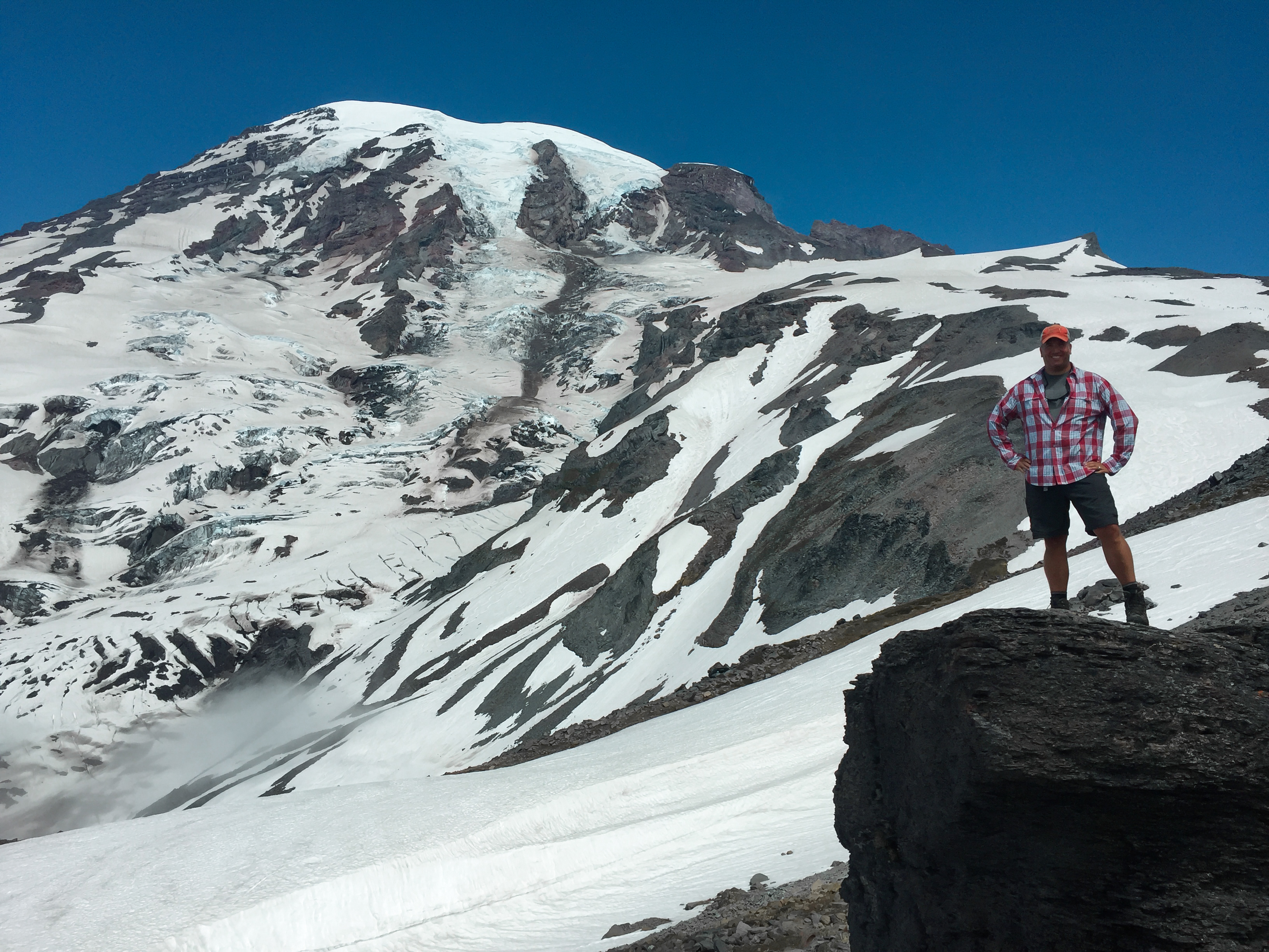 Hiker standing on a rocky outcrop with the massive snow and glacier-covered face of Mount Rainier rising directly behind under a blue sky