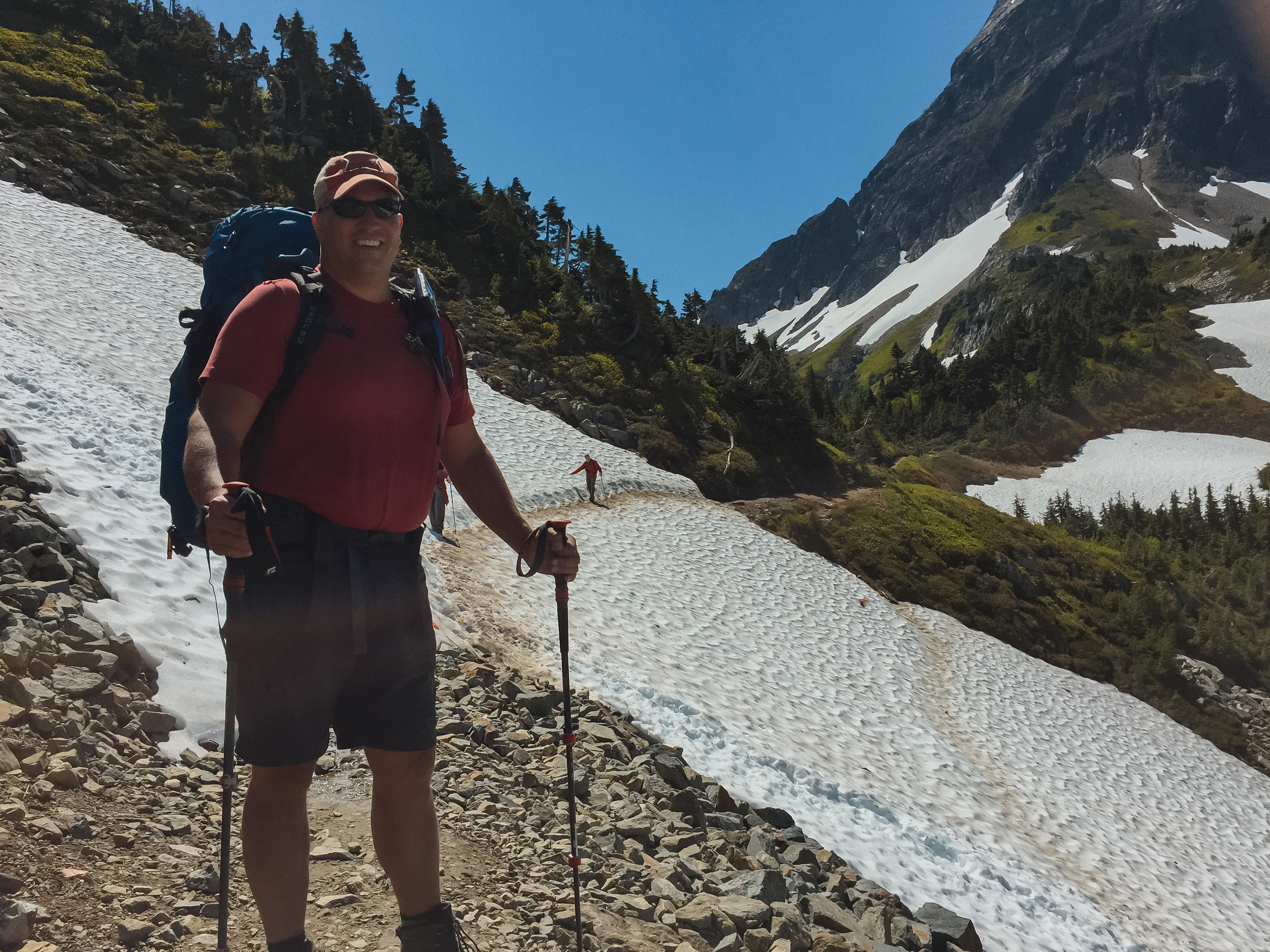 Hiker with full pack and poles crossing a snowfield on the approach to Cascade Pass, Cascade peaks rising behind