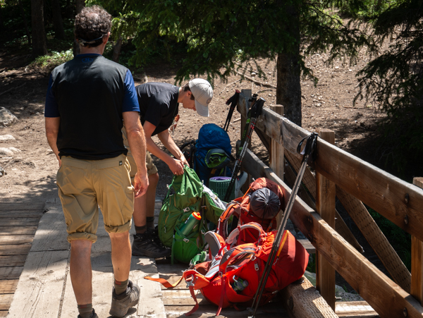 Two hikers pause on a wooden footbridge in a shaded pine forest, adjusting heavy backpacks before continuing up the Lake Fork trail