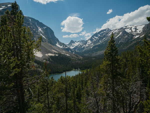 Wide view of the Lake Fork basin with a teal alpine lake nestled among pine forests, snow-capped Beartooth peaks rising in the background under a partly cloudy sky
